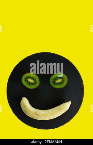 Metà Kiwi e una banana sbucciata su vassoio nero e. sfondo giallo Foto Stock