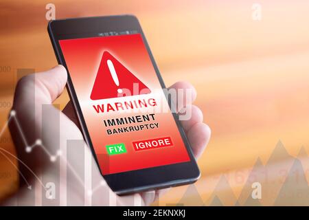 Closeup di mano maschile che tiene moderno telefono cellulare con schermo rosso che mostra il simbolo di avvertimento e la parola con il testo 'IMMINENTE FALLIMENTO'. Foto Stock