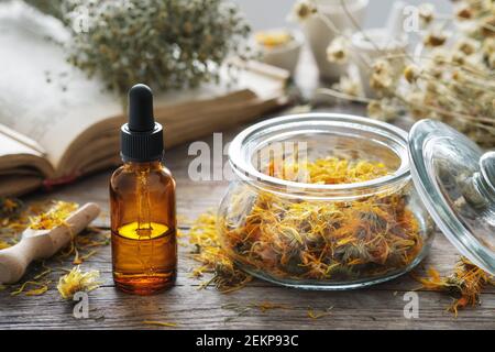 Flacone contagocce di infusione o olio di calendula, vaso di fiori di marigola essiccati, libro di ricette antiche e mazzo di camomilla su sfondo. Medicina alternativa. Foto Stock