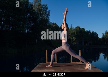 Giovane donna presso il lago che pratica yoga si muove su piattaforma di legno. Bella giovane donna che si esercita in natura, sano stile di vita giovani positivi Foto Stock