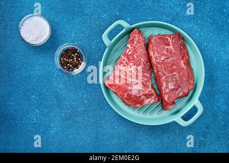 Bistecca cruda su un tagliere con rosmarino e spezie, sfondo blu, vista dall'alto. Carne fresca alla griglia. Bistecca di manzo alla griglia. Foto Stock