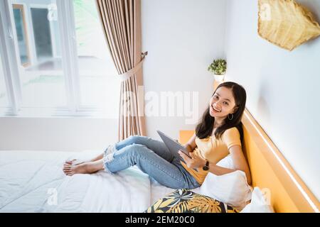 donna asiatica sdraiata a letto con tablet digitale che si tocca dito nella luce solare del mattino Foto Stock