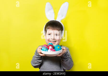 Allegro ragazzo felice con orecchie conigliate sulla testa sta giocando con le uova colorate di Pasqua, battendo le uova di pollo dipinte l'una contro l'altra. Capretto di Pasqua su un grido Foto Stock