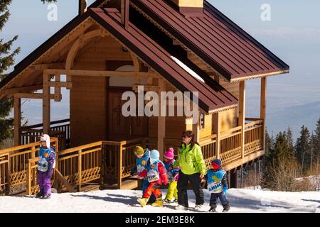 Gruppo di bambini piccoli e istruttore di sci femminile a piedi attraverso la piccola chiesa di legno semplice in inverno nel Monte Vitosha vicino Sofia, Bulgaria Foto Stock