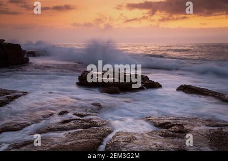 Le onde costiere dell'alba si infrangono sulle rocce e scorrono rapidamente tutte le direzioni per tornare al mare Foto Stock