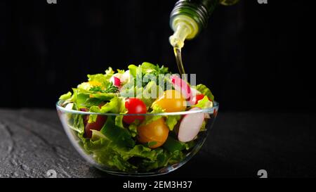 Olio d'oliva versato dalla bottiglia sull'insalata di verdure su nero sfondo Foto Stock