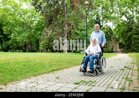 Donna anziana in sedia a rotelle che viene spinta attraverso il parco dal suo uomo, concetto di coppia anziana Foto Stock