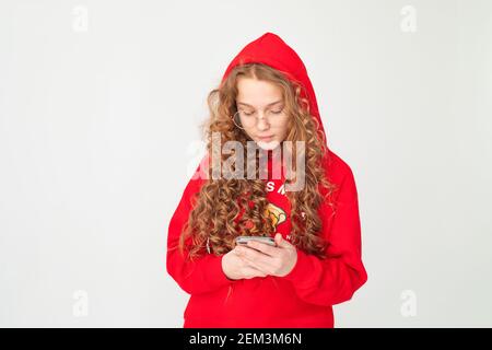 Ragazza adolescente capelli ricci rossi con cappuccio rosso, utilizzando lo smartphone da lettura su sfondo bianco. Concetto di comunicazione online, istruzione. Foto Stock