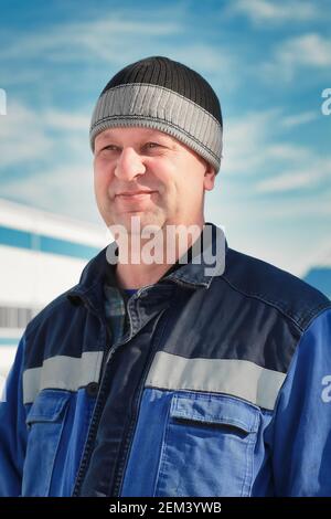 Un uomo in tute e un cappello si trova sulla strada contro il cielo. Ritratto di un vero lavoratore. Derisione Foto Stock