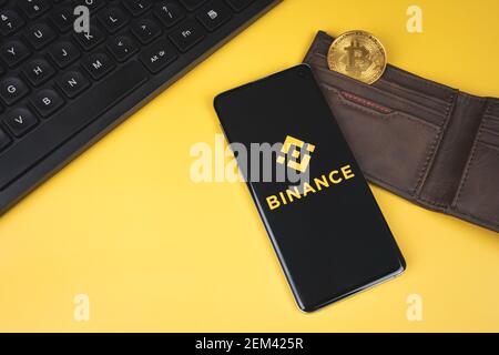 Logo dell'app mobile Binance su uno smartphone, portafoglio con Bitcoin oro e tastiera su sfondo giallo. Foto Stock