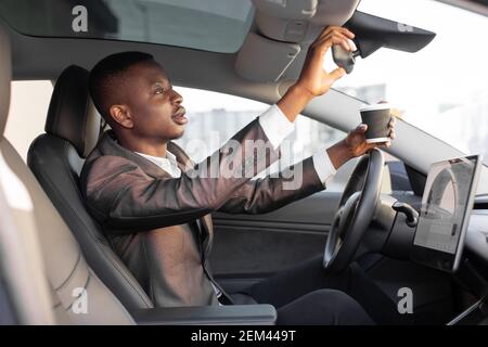 Fai una pausa sulla strada. Bell'uomo africano rilassato in abiti da lavoro, seduto in un moderno veicolo futuristico con display touchscreen, specchio per auto a contatto Foto Stock