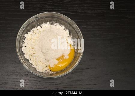 Formaggio di cottage granuloso, uovo, farina in un recipiente di vetro su un tavolo nero primo piano con spazio copia, vista dall'alto Foto Stock