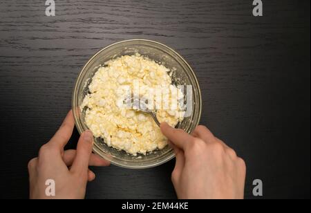 Le mani delle donne mescolano un cucchiaio in una ciotola di vetro di formaggio di cottage granuloso, uovo, farina su un tavolo nero primo piano con spazio di copia, vista dall'alto Foto Stock