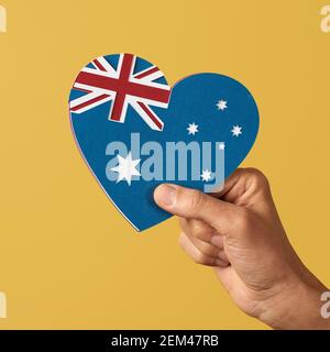 closeup della mano di un uomo che tiene una bandiera australiana a forma di cuore, fatto con tagli di carta di colori diversi, su uno sfondo giallo Foto Stock