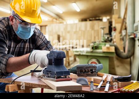 Falegname al lavoro nel laboratorio di carpenteria, indossa casco, occhiali, guanti in pelle e maschera chirurgica per prevenire l'infezione da coronavirus. Eventi Foto Stock