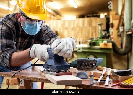 Falegname al lavoro nel laboratorio di carpenteria, indossa casco, occhiali, guanti in pelle e maschera chirurgica per prevenire l'infezione da coronavirus. Eventi Foto Stock