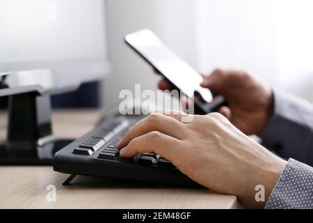 Uomo che usa lo smartphone su sfondo della tastiera del PC. Concetto di comunicazione online, lavoro in ufficio o a casa e pagamento Foto Stock