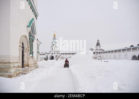 Russia, regione di Mosca, febbraio 2021. UN uomo vestito di calore su un aratro di neve rimuove la neve sul territorio del monastero in una giornata gelida. Foto Stock