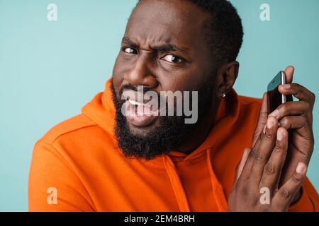 Arrabbiato giovane africano che parla sul telefono cellulare isolato sopra sfondo blu Foto Stock