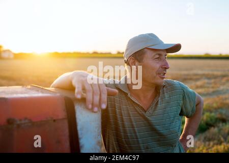 Uomo agronomo anziano in piedi in un campo appoggiato al vecchio trattore dopo la mietitura al tramonto. Foto Stock