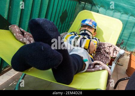 Bambino ragazzo sdraiato su sedia a sdraio nel sole di febbraio mite durante la quarantena casa covid-19, Sopron, Ungheria Foto Stock