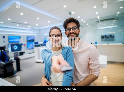 Primo piano ritratto di carino eccitato giovane studente ragazza con occhiali che tengono il telecomando della tv mentre il suo ragazzo abbracciandola in una tecnologia Foto Stock