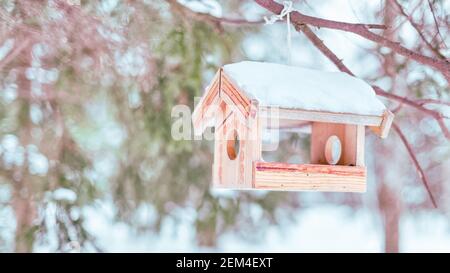 Neve coperta alimentatore di uccelli in legno appeso nella foresta invernale. Immagine chiave alta, messa a fuoco selettiva sull'alimentatore Foto Stock