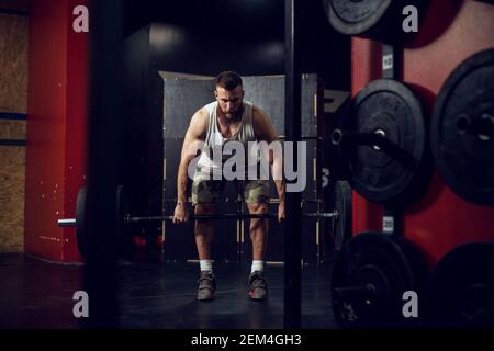 Ritratto di giovane bearded messo a fuoco forte uomo muscolare fitness accovacciato con un pesante barbell davanti mentre fa un deadlift esercizio. Foto Stock