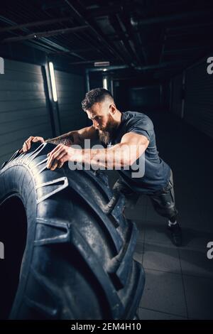 Ritratto primo piano vista di forte muscoloso bearded corto capello bodybuilder uomo che capovolgeva un pneumatico enorme nel garage sotterraneo scuro o palestra urbana. Foto Stock
