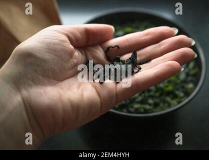 vista ravvicinata delle alghe secche di wakame in mano alla donna con immersione in acqua calda wakame sullo sfondo Foto Stock