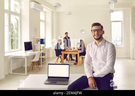 Giovane uomo d'affari seduto sulla scrivania con computer portatile con schermo vuoto in ufficio aziendale Foto Stock