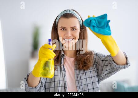 Ritratto di giovane donna sorridente in guanti di gomma per la pulizia del vetro con detergente spray, all'interno Foto Stock