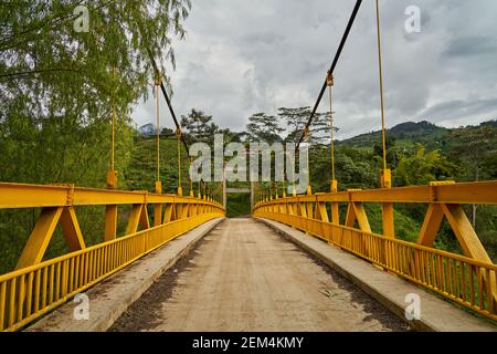 Vecchio ponte sospeso con vernice gialla e un singolo percorso che conduce nella fitta foresta di Jardin in Colombia, Sud America Foto Stock