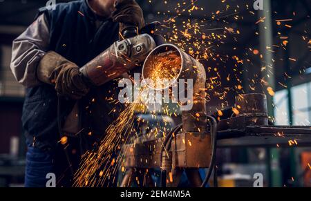 Vista ravvicinata laterale di un lavoratore professionista concentrato lavoro uniforme sulla scultura del tubo metallico con un elettrico macinare mentre le scintille volano in t Foto Stock