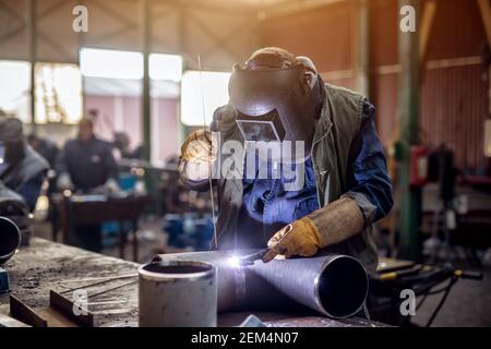 Saldatrice professionale in uniforme protettiva e maschera saldante tubo metallico sul tavolo industriale con altri lavoratori dietro nell'officina industriale. Foto Stock