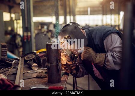 Saldatrice professionale in uniforme protettiva e maschera saldante tubo metallico sul tavolo industriale con altri utensili nell'officina industriale. Foto Stock