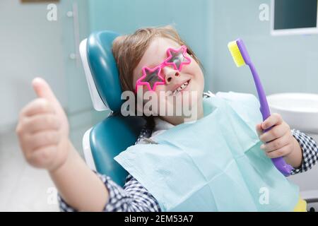 Bel ragazzo che indossa occhiali protettivi, mostrando i pollici e tenendo lo spazzolino in clinica dentale Foto Stock