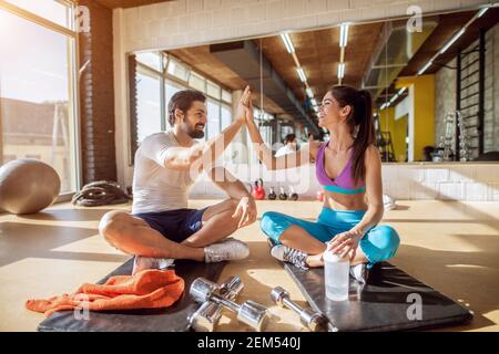 Primo piano di attraente giovane coppia di fitness che si aggrappano le mani insieme mentre si siede sul tappetino nero e ha un fai una pausa in palestra di fronte all'hotel Foto Stock