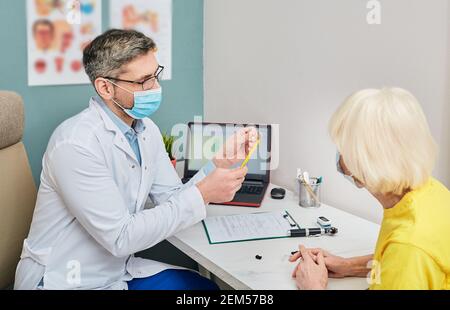 Il medico che indossa una maschera medica mostra un apparecchio acustico ad una donna anziana per il trattamento della sordità durante il coronavirus pandemia Foto Stock