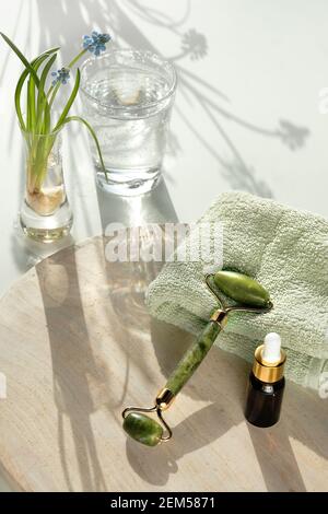 Rullo facciale in pietra di giada verde per il massaggio del viso su asciugamano ripiegato. Bottiglia di vetro scuro con olio essenziale. Pietra bianca e fondo tessile, primavera Foto Stock