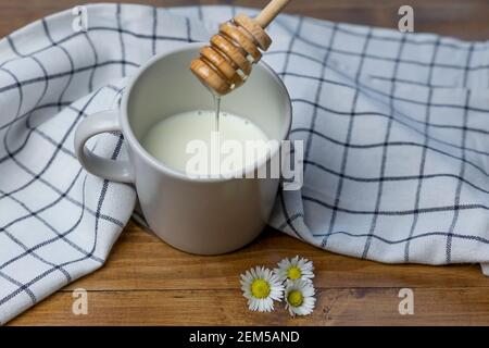 Un cucchiaino di miele versa il miele in una tazza di latte su un tavolo di legno con margherite Foto Stock