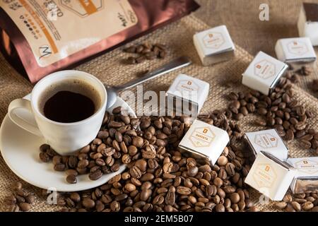 Tiraspol, Moldavia - 23 febbraio 2021: Tazza di caffè, chicchi di caffè e dolci baristi su sackcloth, sullo sfondo una confezione di caffè macinato Bari Foto Stock