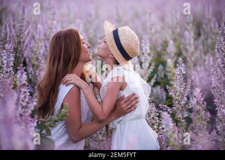 La giovane madre in cialde bianca abbraccia la piccola figlia in cappello di paglia. La ragazza bacia la donna tra il campo di salvia viola in fiore. Cura materna, amore. Vacanza wi Foto Stock