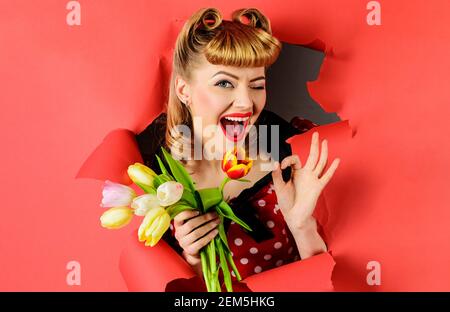 Winking donna con tulipani mostra segno ok. Sorridente Pinup ragazza che guarda attraverso la carta. Donna felice con bouquet di fiori. Foto Stock