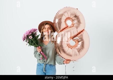 Giovane bella donna che celebra la Giornata Internazionale della Donna, tenendo bouquet di fiori freschi e palloncino in forma di numero otto, sorridente e lookin Foto Stock