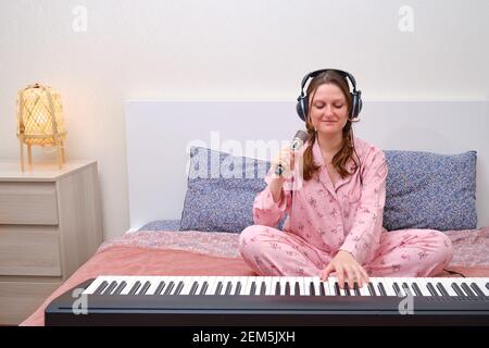 Donna con cuffie canta una canzone in un microfono e. riproduce un sintetizzatore su un letto di casa Foto Stock