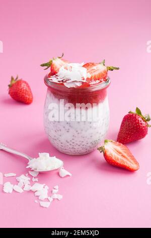 Pudding con chia, fragole e fiocchi di cocco nel vaso di vetro su sfondo rosa. Posizione vetica. Spazio di copia. Foto Stock