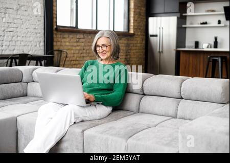 Affascinante anziana donna trascorre il tempo libero in reti sul computer, messaggi di testo, guardare film. La signora matura lavora a distanza con un computer portatile seduto sul divano in un accogliente soggiorno Foto Stock