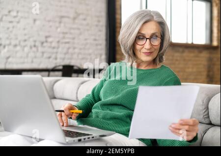 Una donna d'affari anziana sicura sta facendo i documenti seduti con un laptop sul divano a casa, un dipendente senior concentrato che prepara i documenti, controlla le fatture lavorando a casa da remoto Foto Stock