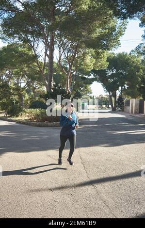 Donna fitness matura, tra i 30 e i 50 anni, in corsa, in viaggio, circondata da alberi di pino, indossare occhiali sportivi, cuffie bluetooth, blu Foto Stock